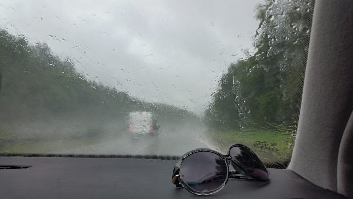 Eine Sonnenbrille liegt auf der Ablage eines Autos auf der Beifahrerseite, die Windschutzscheibe ist voller Regenwasser.