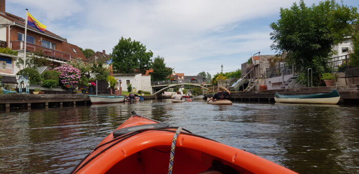 Das Bild zeigt die Friedrichstädter Altstadt von meinem Leih-Kajak aus. Rechts und links der Gracht liegen die Boote der Anwohner, an den Anlegern sind Blumen gepflanzt und die Fahne Nordfrieslands weht in einem der Gärten.