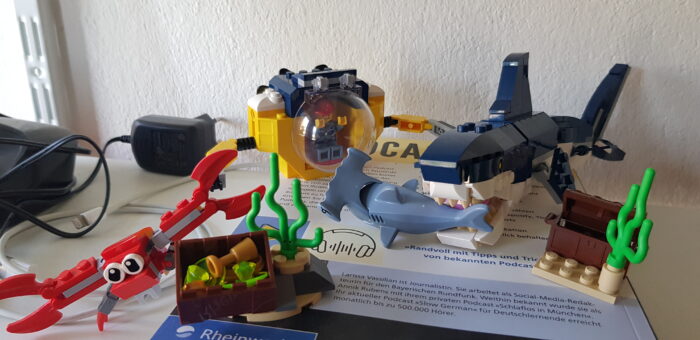 Das Bild zeigt zwei miteinander kombinierte Legosets. In der dargestellten Szene beobachtet ein Mensch in einem Forschungsuboot, wie ein Megalodon einen Hammerhai frisst, während im Vordergrund ein riesiger Krebs die Flucht ergreift.