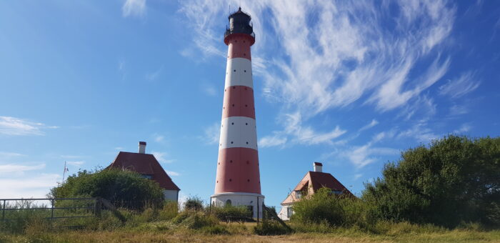 Das Bild zeigt die Leutturmwarft von Westerhever, zu stehen ist der berühmte Leuchtturm mit seinen beiden Nebenhäusern. Die Sonne scheint und am Himmel sind nur einige Cirruswolken zu sehen.