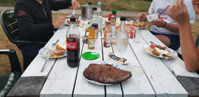 Das Bild zeigt einen Tisch im Landhaus-Stil auf dem Getränke, Grillsaucen und diverses Essen steht. Im Vordergrund steht ein Teller, der komplett von einem Stück Flanksteak eingenommen wird.