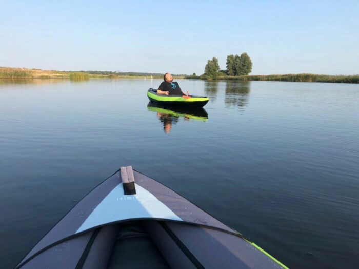 Im Vordergrund ist die Spitze von Gesches Kayak zu sehen, in der Bildmitte im Hintergrund lehne ich mich in meinem Kayal zurück und genieße die Abendsonne auf der Treene. Der Wasseroberfläche ist fast völlig glatt.