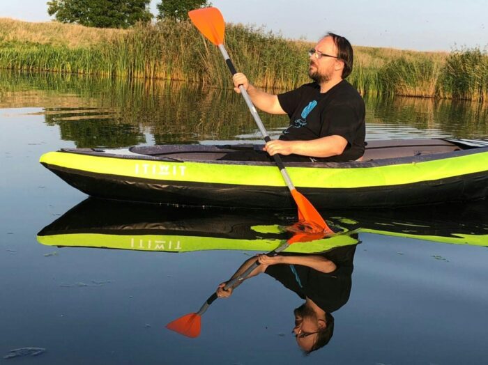 Das Bild zeigt mich, wie ich in meinem Kayak von rechts nach links an der Kamera vorbeipaddele, ein Paddelblatt ist dabei im Wasser, eines in der Luft. Das Wasser ist so glatt, dass ich mich darin spiegele.